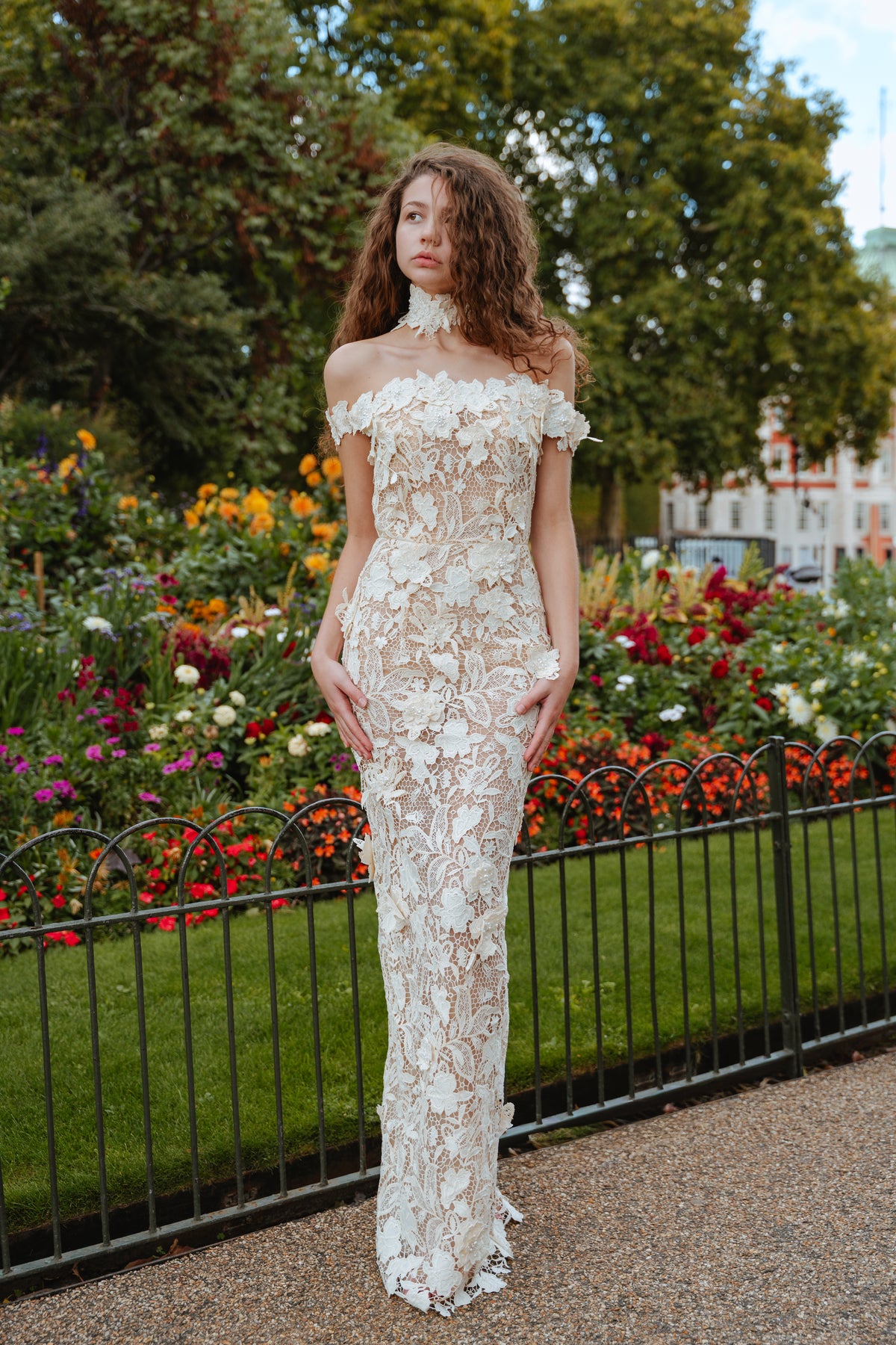 Model wearing an ivory off-the-shoulder gown with floral appliques beside a flower garden.